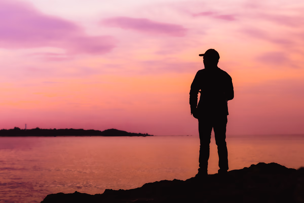 Silhouetted figure on rocky shore gazing at a pink and purple sunset over the se