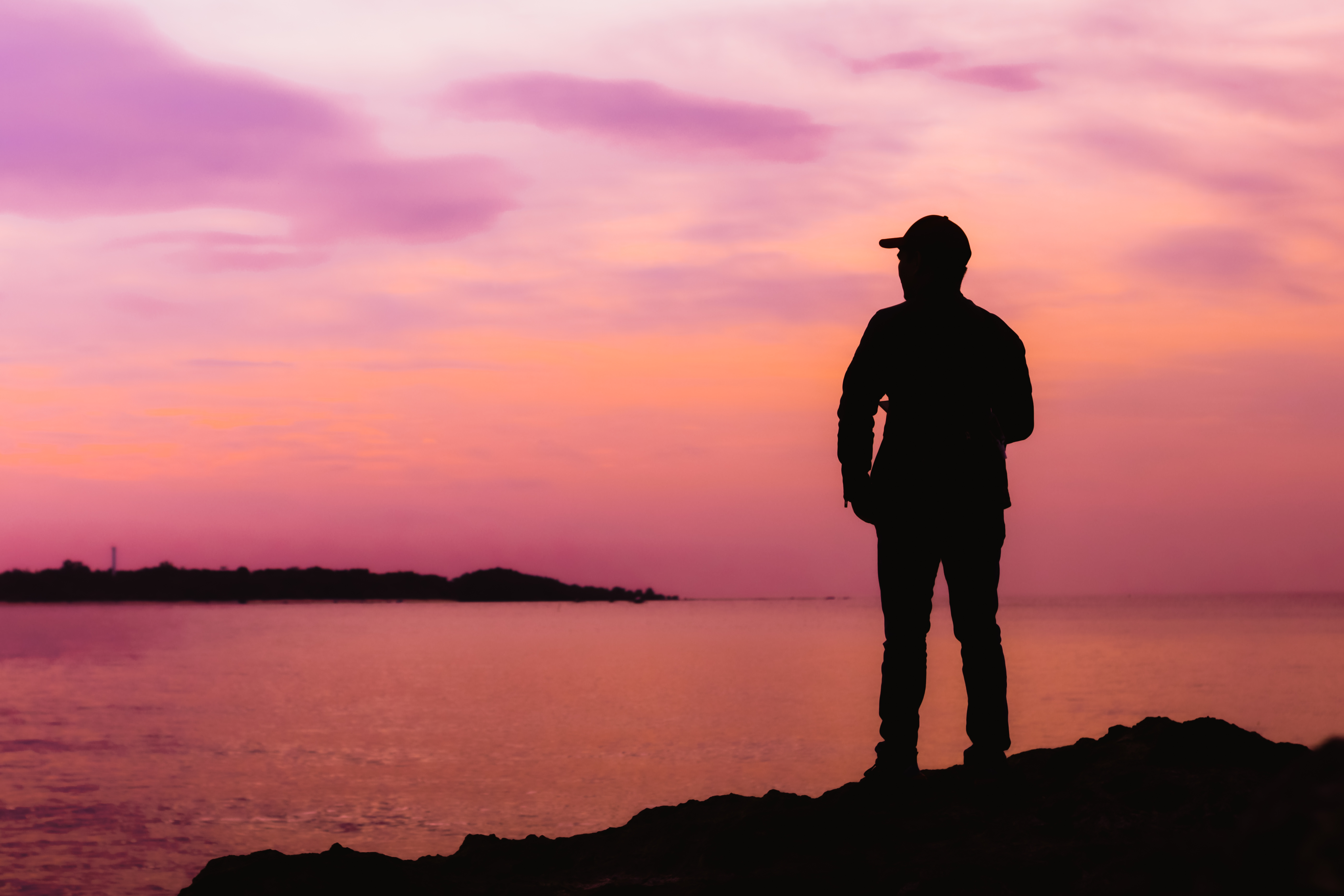 Silhouetted figure on rocky shore gazing at a pink and purple sunset over the sea