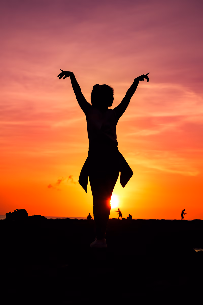 Silhouette of a woman dancing on rocky shore against a purple and orange sunset